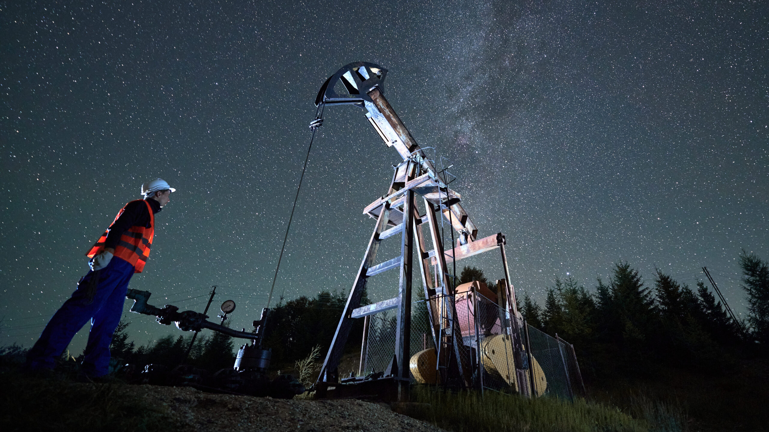 Bottom view of man in workwear standing to the left of oil pump and watching for its activities. Mechanic controls efficient night work of oil drilling rig. Starry sky and forest on background.